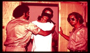 Blindfolded and bound prisoner standing with two prison guards wearing sunglasses.