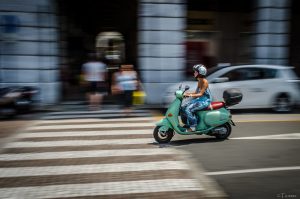 A woman wearing a helmet driving a Vespa motor scooter while pedestrians walk nearby.