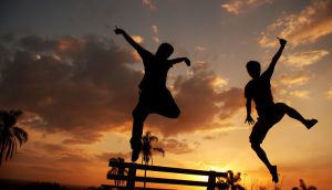 Two people jumping up with hands and feet in the air near a bench at sunrise or sunset.