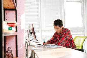 Picture of young african student writing notes in notebook while sitting near computer