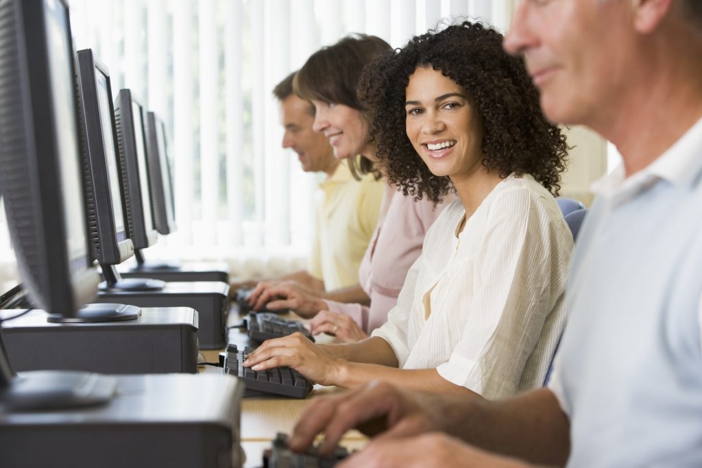 Woman sitting in the middle of people staring at laptops; she is turned toward the viewer smiling.