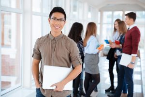 Portrait of a cheerful male student standing in university hall with classmates on background